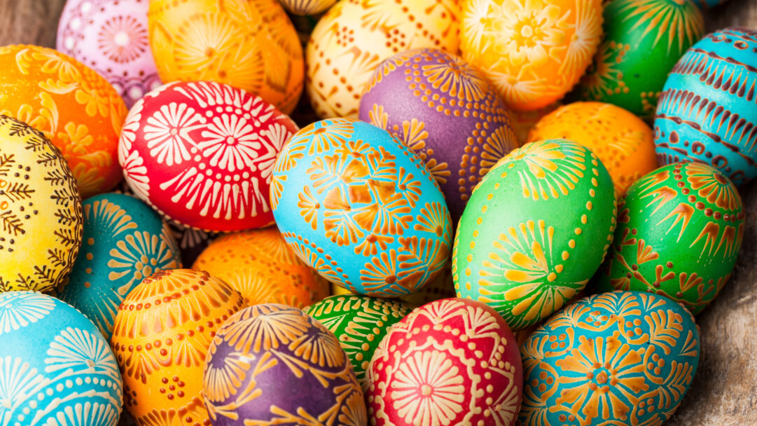 A vibrant 4K wallpaper showcasing a close-up of numerous traditional Colorful Lithuanian Easter Eggs, piled together in a festive arrangement. Each egg displays intricate wax-resist patterns in contrasting shades like golden swirls on deep red, teal, green, and purple, radiating a joyful and artistic spring spirit.
