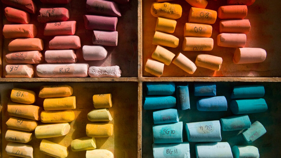 A vibrant 4K wallpaper showcasing an array of colorful pastel chalk art supplies neatly arranged within a partitioned wooden box. The distinct blocks of warm reds, vibrant yellows, and cool blues and greens are accentuated by soft light, highlighting their powdery textures and creating a rich, artistic ambiance.