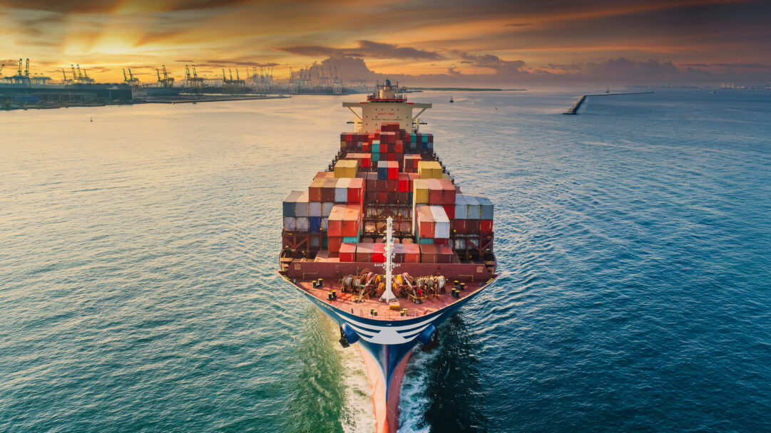A breathtaking 4K wallpaper showcasing a massive container ship, loaded with diverse cargo containers, approaching a bustling commercial port in Thailand as the sun begins to set. The vibrant orange and purple hues of the sunset sky dramatically illuminate the ship's path, casting a golden shimmer across the tranquil ocean waters and silhouetting the numerous cranes of the distant port.