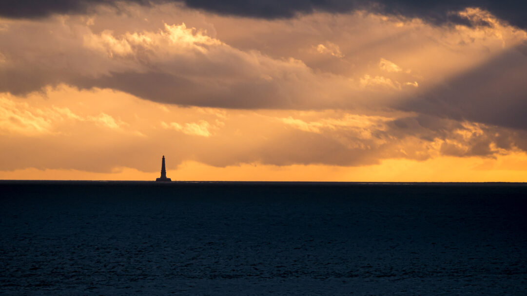 An evocative 4K wallpaper capturing the Cordouan Lighthouse as a striking silhouette against a sunset over the Atlantic Ocean off Royan, France. The vast dark expanse of the ocean below contrasts sharply with the radiant, golden-orange sky, where dramatic clouds catch the last rays of light and cast deep shadows, creating a serene yet powerful mood.