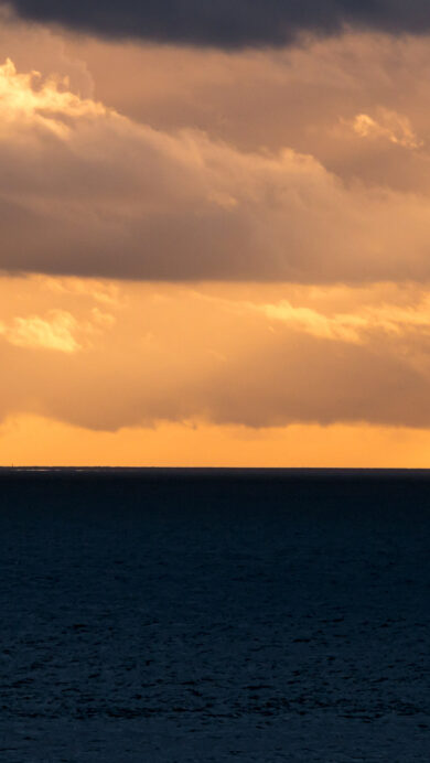 An evocative 4K wallpaper capturing the Cordouan Lighthouse as a striking silhouette against a sunset over the Atlantic Ocean off Royan, France. The vast dark expanse of the ocean below contrasts sharply with the radiant, golden-orange sky, where dramatic clouds catch the last rays of light and cast deep shadows, creating a serene yet powerful mood.