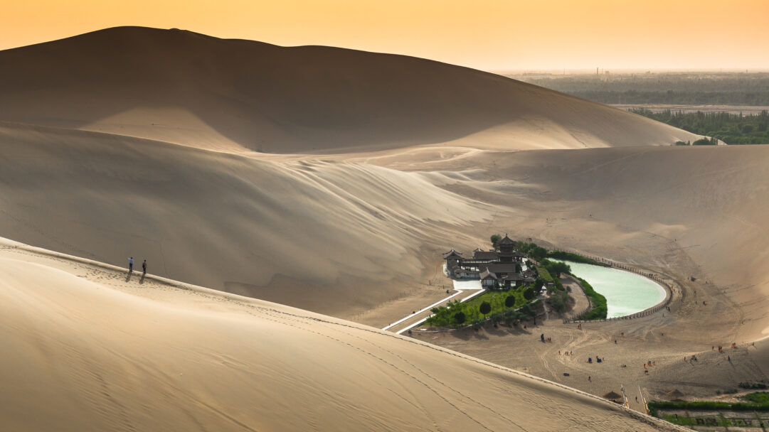 A breathtaking 4K wallpaper captures the iconic Crescent Lake and its traditional Chinese buildings, a thriving desert oasis nestled within the vast, golden sand dunes of Dunhuang, Gansu, China, with tiny figures traversing the slopes. The luminous crescent-shaped lake and lush green vegetation dramatically contrast with the sweeping beige dunes, all bathed in the soft, warm glow of the sky, creating a serene and awe-inspiring landscape.