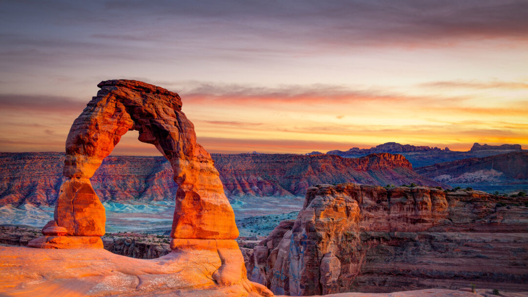 A magnificent 4K wallpaper capturing Delicate Arch, a grand natural sandstone arch, standing proudly amidst the red rock landscape of Arches National Park in Utah. The sunset paints the sky with fiery oranges and soft purples, casting a vibrant, warm glow that brilliantly illuminates the arch itself and the distant mesas.