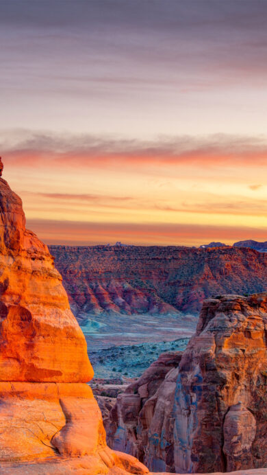 A magnificent 4K wallpaper capturing Delicate Arch, a grand natural sandstone arch, standing proudly amidst the red rock landscape of Arches National Park in Utah. The sunset paints the sky with fiery oranges and soft purples, casting a vibrant, warm glow that brilliantly illuminates the arch itself and the distant mesas.