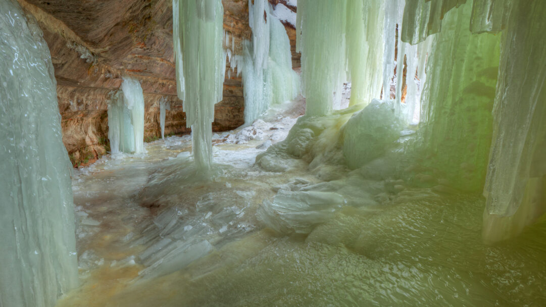 A captivating 4K wallpaper transports you to the Eben Ice Caves of Michigan's Upper Peninsula, revealing a surreal landscape of immense frozen waterfalls and ice-covered cavern floors. The towering, pale green and blue ice formations glow with an ethereal light, creating a serene and magical frozen spectacle.