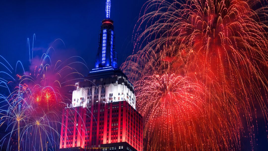 A dazzling 4K wallpaper features the Empire State Building illuminated in patriotic colors amidst a Fourth of July fireworks display over New York City. The building's red, white, and blue lighting harmonizes with the expansive red and blue fireworks exploding across the dark night sky, conveying a celebratory and grand atmosphere.