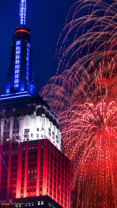A dazzling 4K wallpaper features the Empire State Building illuminated in patriotic colors amidst a Fourth of July fireworks display over New York City. The building's red, white, and blue lighting harmonizes with the expansive red and blue fireworks exploding across the dark night sky, conveying a celebratory and grand atmosphere.