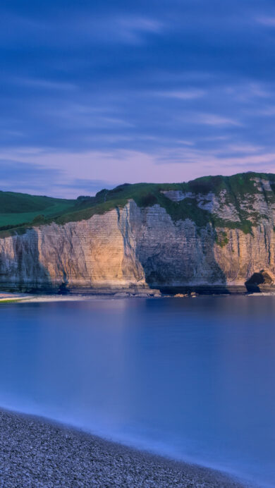 A breathtaking 4K wallpaper showcasing the iconic Étretat Chalk Cliffs in Normandy, France, nestled along a serene pebble beach at twilight. Warm artificial lights illuminate parts of the majestic white cliffs, contrasting beautifully with the cool, calm seascape where the sky transitions from deep blue to a soft, ethereal pink at the horizon.