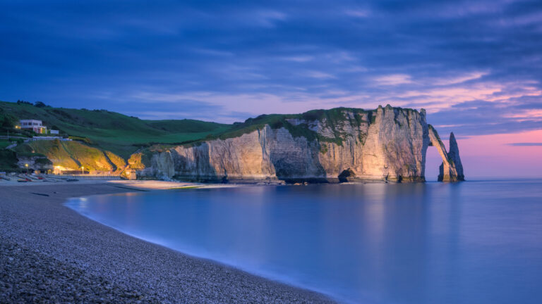 A breathtaking 4K wallpaper showcasing the iconic Étretat Chalk Cliffs in Normandy, France, nestled along a serene pebble beach at twilight. Warm artificial lights illuminate parts of the majestic white cliffs, contrasting beautifully with the cool, calm seascape where the sky transitions from deep blue to a soft, ethereal pink at the horizon.