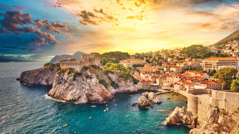 A dramatic 4K wallpaper showcasing Fort Lovrijenac standing majestically on a rocky outcrop in Dubrovnik's West Harbor, with the historic city sprawling along the Croatian coastline. The intense sunset bathes the scene in a gradient of golden light and deep blue hues, illuminating the fortress walls and the tranquil water where kayakers paddle.