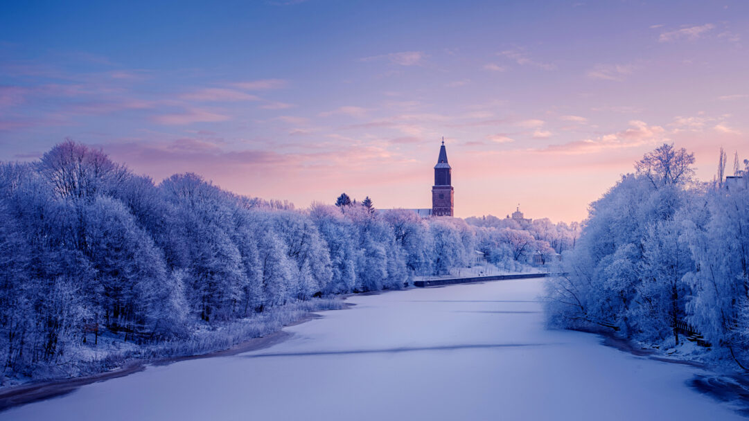 An atmospheric 4K wallpaper of the frozen Aura River winding through a snow-covered winter landscape in Turku, Finland, with the prominent Cathedral tower rising above frosted trees. The soft pastels of the dawn sky blend seamlessly from deep blue to delicate pink and purple, casting an ethereal glow over the brilliant white, frost-laden trees and the smooth, icy river surface.