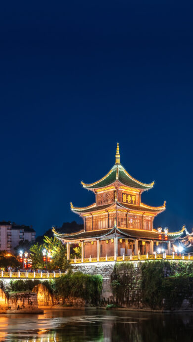 An enchanting 4K wallpaper showcasing the historic Guiyang Jiaxiu Tower, brilliantly lit against the night sky, standing gracefully on its foundation in a river surrounded by illuminated bridges and city architecture. A prominent, luminous full moon hangs in the deep blue sky, its warm light reflecting softly on the dark waters and enhancing the tower's traditional golden and green hues.