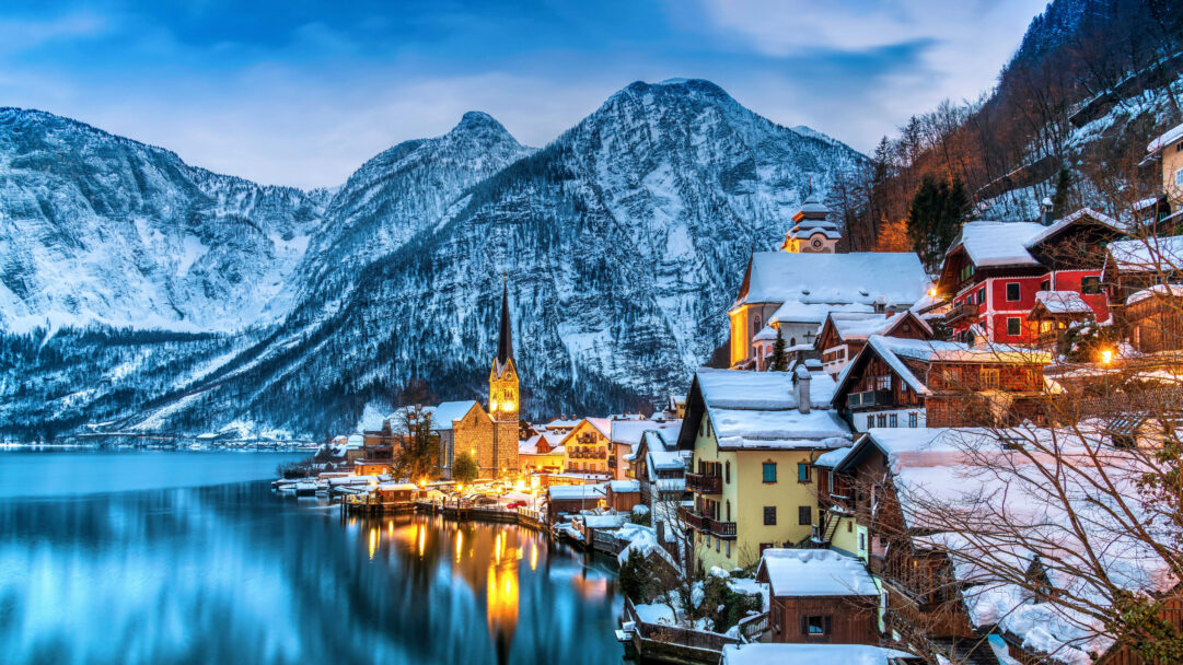 A breathtaking 4K wallpaper featuring Hallstatt, Austria, a picturesque winter village nestled beside a serene lake and dramatic snow-capped mountains. The warm glow of lights from the village buildings and the prominent church steeple reflects beautifully on the tranquil lake, creating a magical contrast with the cool blue twilight and snow.