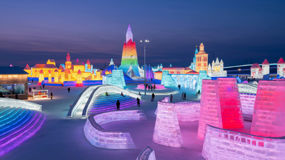 A captivating 4K wallpaper reveals the magical Harbin International Ice and Snow Sculpture Festival in Harbin, China, set against a dark night sky. Brilliantly illuminated ice sculptures glow with electric blues, fiery reds, and warm yellows, creating a fantastical, festive atmosphere as visitors explore the icy wonderland.