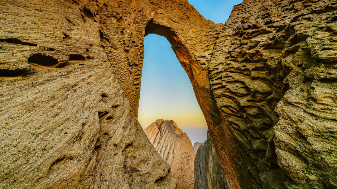 An epic 4K wallpaper captures Heaven's Gate Cave, a monumental natural rock arch within Tianmen Mountain, China. The dramatic, textured golden-brown rock formations frame a distant peak against a serene, gradient sky, evoking a profound sense of natural wonder and timelessness.