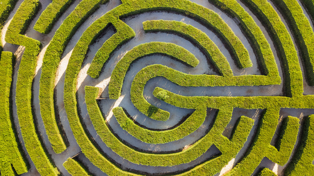 A captivating 4K wallpaper showcasing an aerial view of the winding Hedge Maze within Cyherbia Botanical Park in Cyprus. The perfectly manicured, vibrant green hedges sharply contrast with the sun-drenched gravel paths, creating an intricate and inviting pattern of light and shadow.