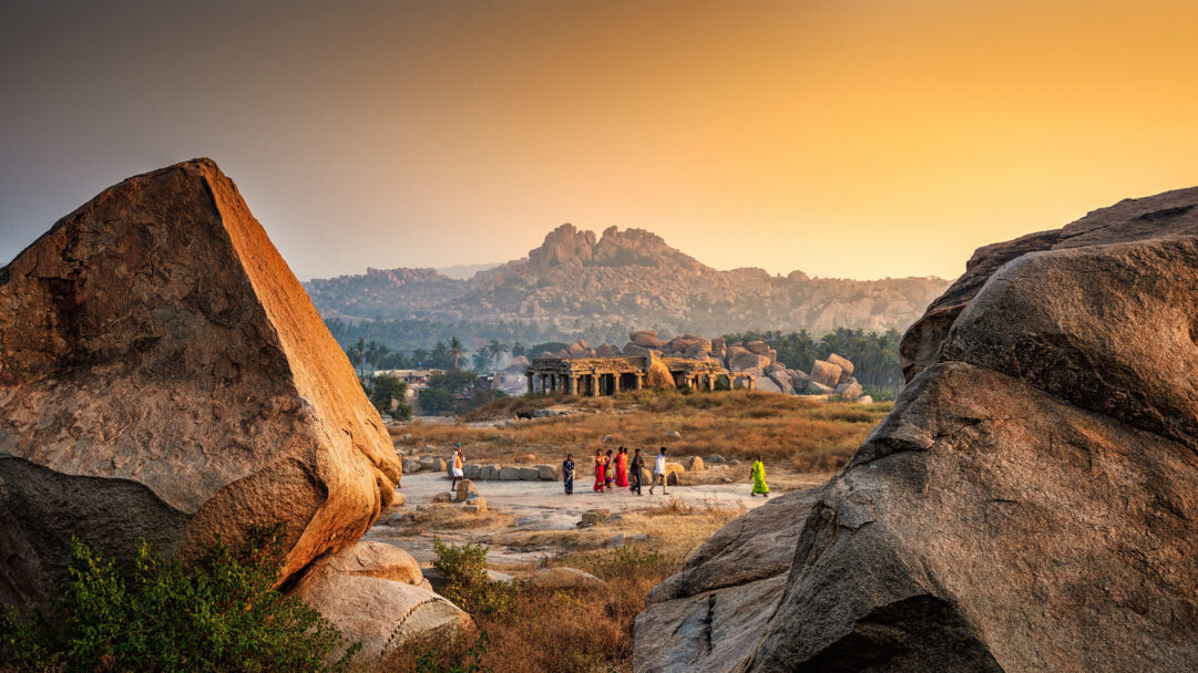 An ancient 4K wallpaper features the iconic Hemakuta Hill Temples in Hampi, Karnataka, India, nestled among colossal boulders under a vast sky. The warm, golden glow of the sunset casts long shadows, illuminating the rugged textures of the ancient stone structures and creating a majestic, timeless atmosphere as visitors explore the historic landscape.