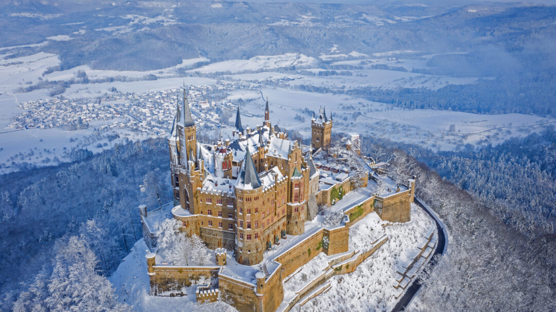 A majestic 4K wallpaper featuring Hohenzollern Castle perched atop a snow-covered peak in the German winter landscape. The historic castle’s warm stone towers and roofs are blanketed in pristine white snow, standing out against the expansive, frosty forests and distant valleys below, evoking a serene and timeless winter wonderland.
