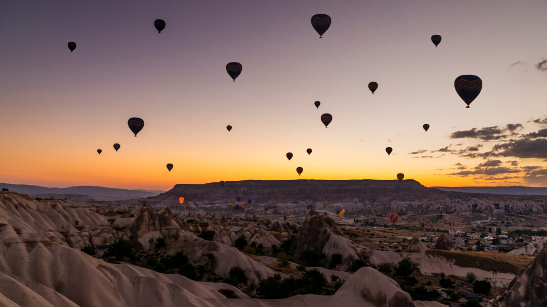 A breathtaking 4K wallpaper capturing numerous hot air balloons gracefully ascending over the unique, undulating landscapes of Cappadocia Valley in Türkiye. The sky, transitioning from a warm golden orange to soft lavender, dramatically silhouettes the distant mountains and the iconic fairy chimney formations, creating an ethereal and serene atmosphere.