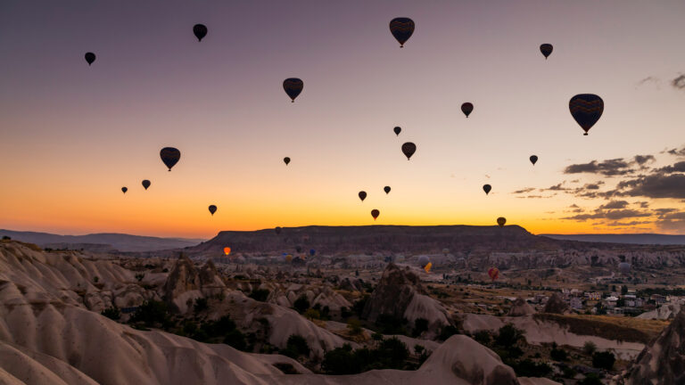 A breathtaking 4K wallpaper capturing numerous hot air balloons gracefully ascending over the unique, undulating landscapes of Cappadocia Valley in Türkiye. The sky, transitioning from a warm golden orange to soft lavender, dramatically silhouettes the distant mountains and the iconic fairy chimney formations, creating an ethereal and serene atmosphere.
