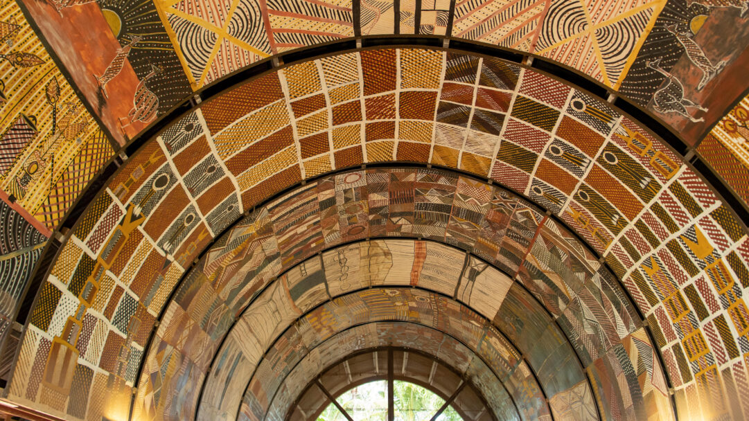 A stunning 4K wallpaper of an expansive arched ceiling adorned with intricate Indigenous Aboriginal art patterns. The diverse collection of abstract designs, featuring dot paintings and cross-hatching in earthy ochre, red, yellow, and black, creates a vibrant, culturally rich tapestry overhead.