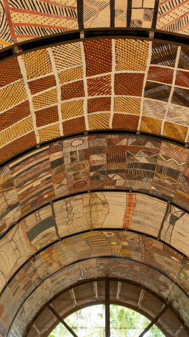 A stunning 4K wallpaper of an expansive arched ceiling adorned with intricate Indigenous Aboriginal art patterns. The diverse collection of abstract designs, featuring dot paintings and cross-hatching in earthy ochre, red, yellow, and black, creates a vibrant, culturally rich tapestry overhead.