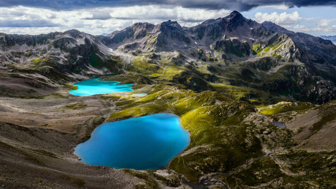 A majestic 4K wallpaper depicts the Jöriseen Lakes as part of the grand mountain landscape of the Silvretta Alps in Switzerland. Its vibrant, glacial blue waters gleam brilliantly, creating a stunning contrast against the rugged peaks, shadowed valleys, and patches of sun-drenched green under a dramatic cloudy sky.