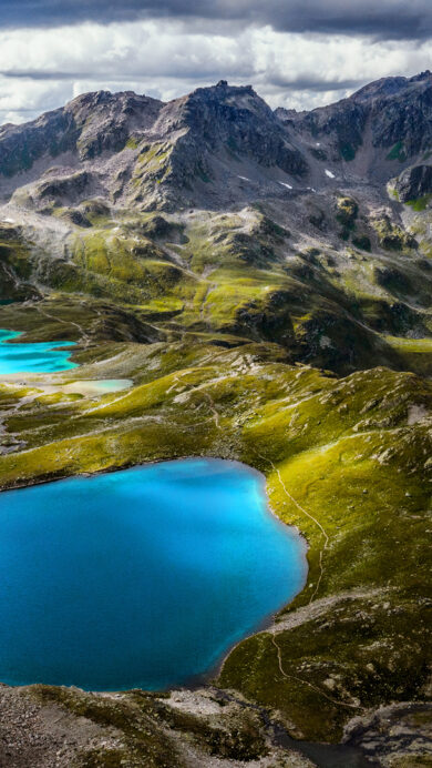 A majestic 4K wallpaper depicts the Jöriseen Lakes as part of the grand mountain landscape of the Silvretta Alps in Switzerland. Its vibrant, glacial blue waters gleam brilliantly, creating a stunning contrast against the rugged peaks, shadowed valleys, and patches of sun-drenched green under a dramatic cloudy sky.