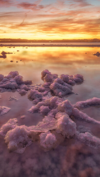 A magical 4K wallpaper of the Laguna de Torrevieja Pink Salt Lake at sunset in Alicante, Spain. The vivid pink waters and crystallized salt formations reflect the fiery orange, yellow, and deep purple hues of the dramatic sky, creating an ethereal and serene landscape.