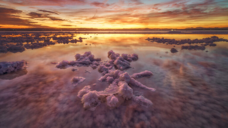 A magical 4K wallpaper of the Laguna de Torrevieja Pink Salt Lake at sunset in Alicante, Spain. The vivid pink waters and crystallized salt formations reflect the fiery orange, yellow, and deep purple hues of the dramatic sky, creating an ethereal and serene landscape.