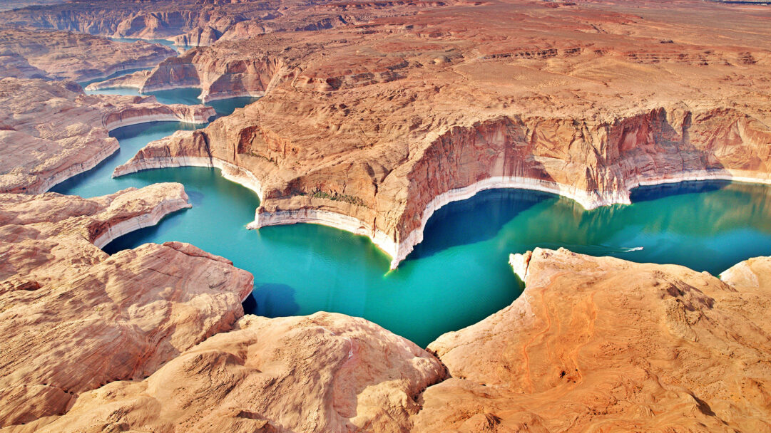 An expansive 4K wallpaper capturing the aerial view of Lake Powell, where the vibrant turquoise Colorado River dramatically winds through vast, stratified sandstone canyons. The striking contrast between the brilliant water and the rugged, sun-baked walls, highlighted by pale erosion lines, creates a majestic and serene natural spectacle.