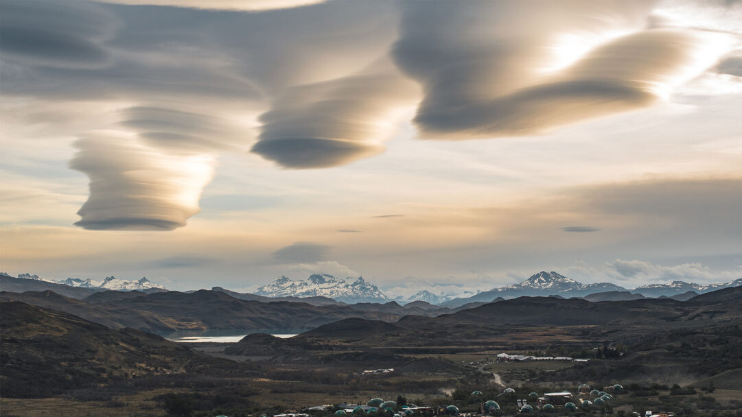 An otherworldly 4K wallpaper showcasing fascinating lenticular clouds above the vast Patagonia mountain landscape. These surreal, stacked clouds, illuminated by soft golden light, create a striking contrast with the shadowed valleys and rugged, snow-capped peaks stretching into the distance.