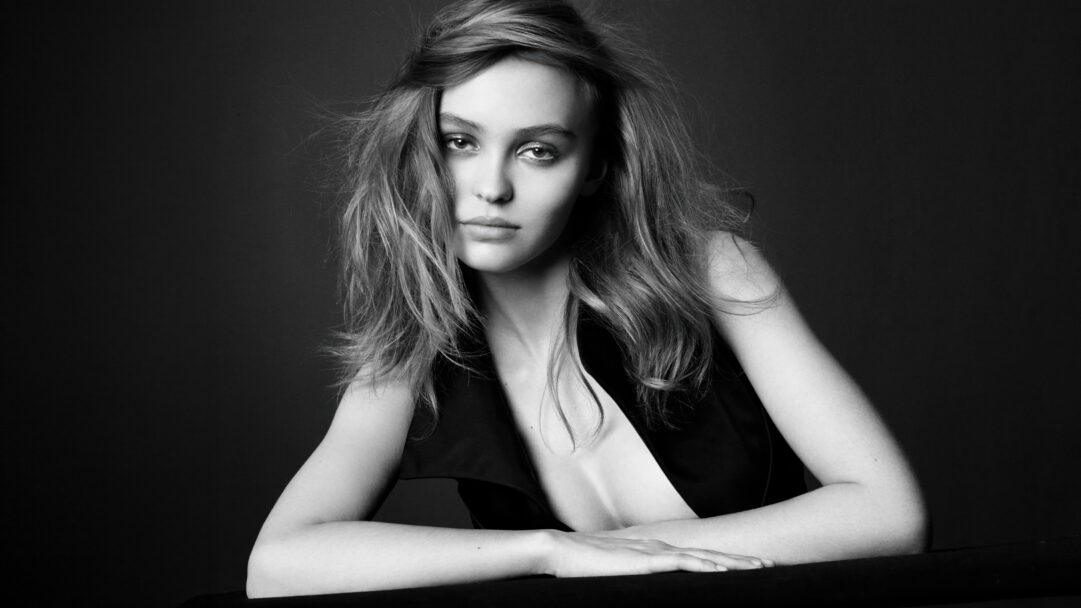 An intimate 4K wallpaper capturing a black and white portrait of Lily Rose Depp leaning forward with a dark, minimalist background. Her intense, direct gaze and subtly windblown hair are illuminated by soft lighting, emphasizing the dramatic contrast of the monochrome palette.