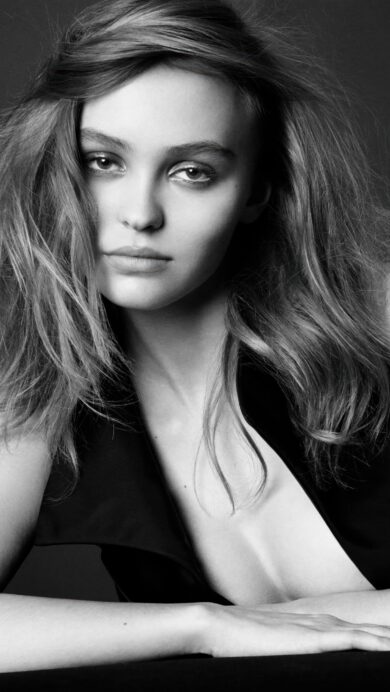 An intimate 4K wallpaper capturing a black and white portrait of Lily Rose Depp leaning forward with a dark, minimalist background. Her intense, direct gaze and subtly windblown hair are illuminated by soft lighting, emphasizing the dramatic contrast of the monochrome palette.