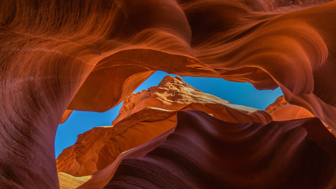 A breathtaking 4K wallpaper captures the majestic sandstone formations of Lower Antelope Canyon in Page, Arizona. The vibrant interplay of warm oranges and deep reds across the sculpted, wave-like sandstone walls creates a mesmerizing dance of light and shadow against the brilliant sliver of blue sky above.