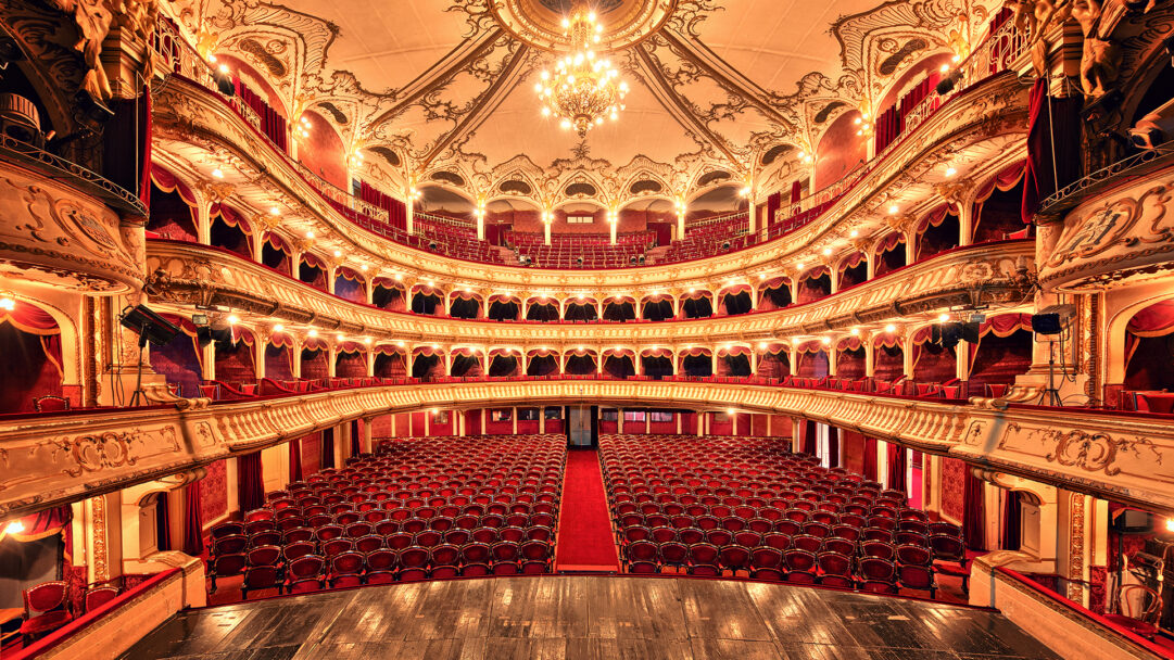 A magnificent 4K wallpaper captures the opulent interior of the Lucian Blaga National Theater auditorium in Cluj-Napoca, Romania. Its multiple tiers of gilded balconies, adorned with intricate rococo designs, sweep around a vast expanse of empty crimson velvet seating, all bathed in the warm glow of countless chandeliers and accent lights.