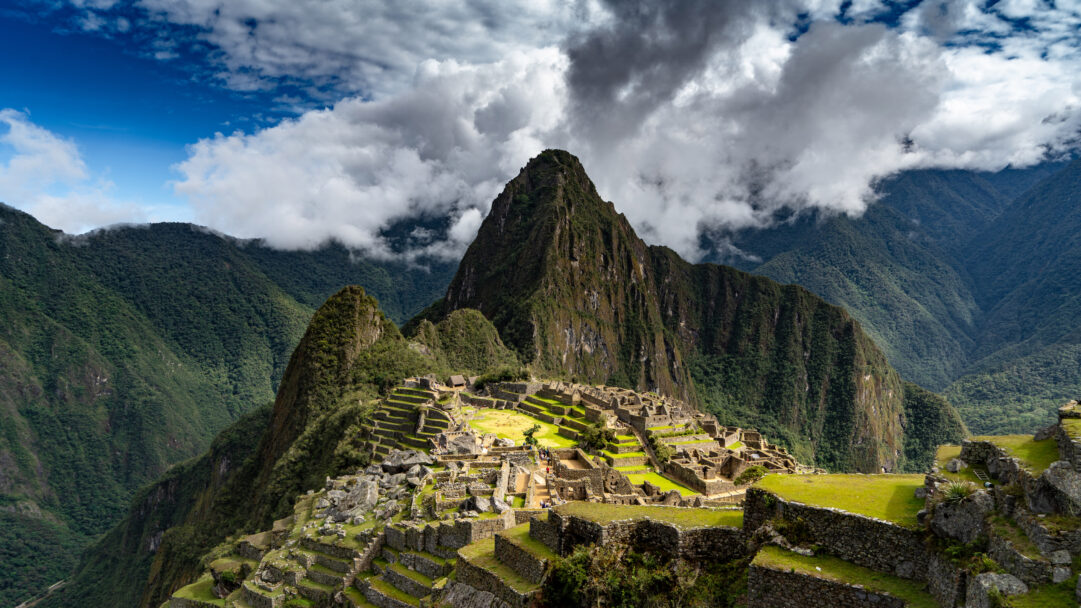 A breathtaking 4K wallpaper of the ancient city of Machu Picchu, nestled high in the Peruvian Andes Mountains. Dramatic clouds partially obscure the towering, jungle-clad peaks, while sunlight highlights the intricate stonework and verdant terraces of the ancient city, evoking a sense of timeless wonder.