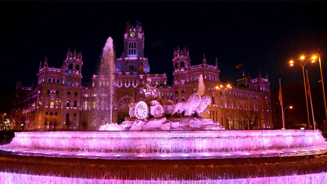 A magnificent 4K wallpaper featuring Madrid's Cibeles Fountain and City Hall brilliantly illuminated in pink at night for International Women's Day. The striking pink light dramatically highlights the historic architecture and the gushing water, creating a vibrant and celebratory urban landscape.