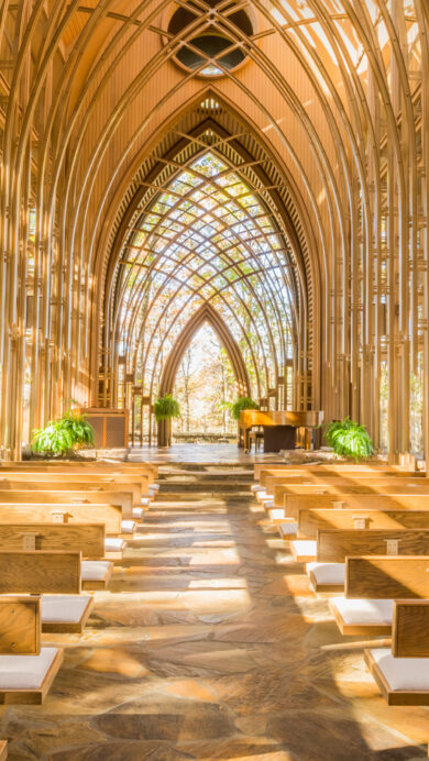 A radiant 4K wallpaper showcasing the interior of the Mildred B. Cooper Memorial Chapel in Bella Vista, Arkansas, revealing its unique architectural design integrated with the surrounding natural environment. Warm sunlight streams through the intricate latticework of wooden arches and glass walls, casting beautiful patterns on the flagstone floor and wooden pews, creating a luminous and tranquil atmosphere amidst the autumn foliage.