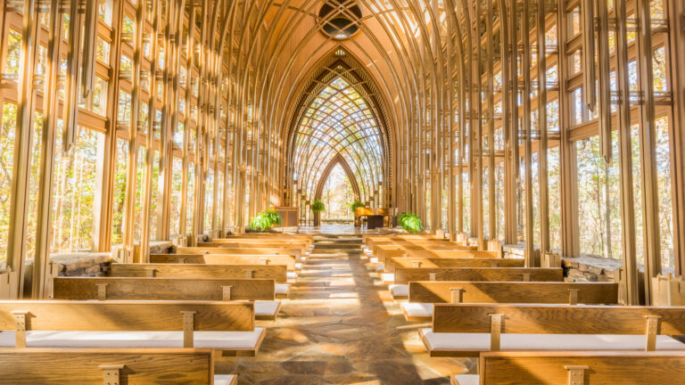 A radiant 4K wallpaper showcasing the interior of the Mildred B. Cooper Memorial Chapel in Bella Vista, Arkansas, revealing its unique architectural design integrated with the surrounding natural environment. Warm sunlight streams through the intricate latticework of wooden arches and glass walls, casting beautiful patterns on the flagstone floor and wooden pews, creating a luminous and tranquil atmosphere amidst the autumn foliage.