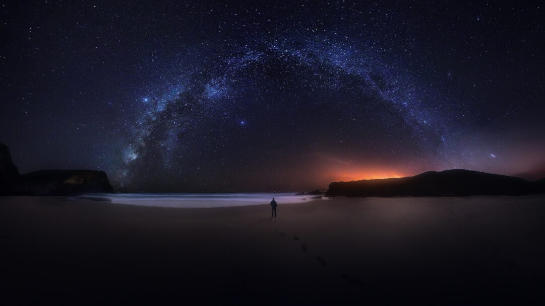 A mesmerizing 4K wallpaper unveils the Milky Way night sky spanning gracefully over a secluded beach on the Southwest Alentejo Portugal coast, with a lone figure gazing out at the ocean. The vibrant celestial band of countless stars dramatically arches across the deep blue-black heavens, contrasting with the warm, distant orange glow on the horizon and the reflective wet sand underfoot.