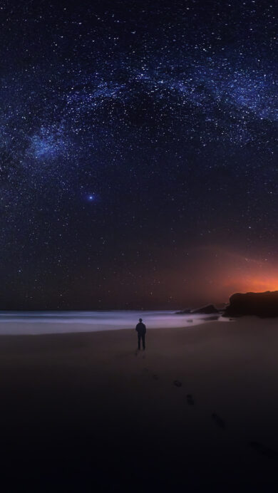 A mesmerizing 4K wallpaper unveils the Milky Way night sky spanning gracefully over a secluded beach on the Southwest Alentejo Portugal coast, with a lone figure gazing out at the ocean. The vibrant celestial band of countless stars dramatically arches across the deep blue-black heavens, contrasting with the warm, distant orange glow on the horizon and the reflective wet sand underfoot.