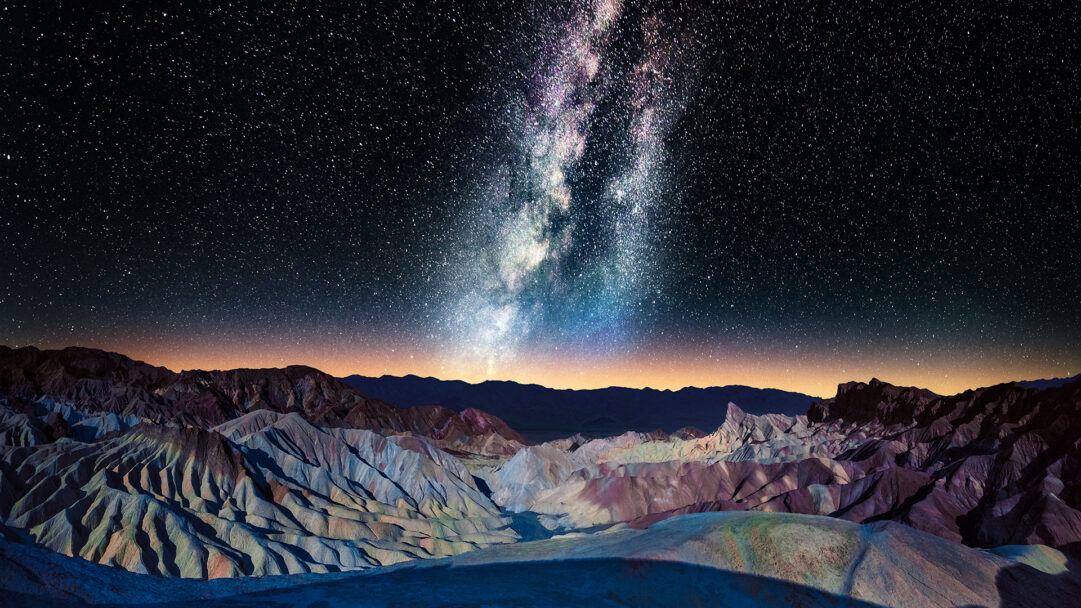 A breathtaking 4K wallpaper featuring the luminous Milky Way arching majestically over the dramatic, multi-hued badlands of Zabriskie Point in Death Valley, California. The celestial river of stars brilliantly contrasts with the subtly illuminated, textured landscape below, creating a truly awe-inspiring nocturnal vista.