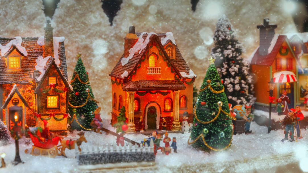 A delightful 4K wallpaper presents a charming miniature Christmas holiday village scene, reminiscent of Strasbourg, France, blanketed in pristine snow. Warm golden light spills from the windows of several ornate, snow-covered houses, accentuating the festive atmosphere created by tiny decorated Christmas trees and playful village figures, all softly framed by a bokeh of gently falling snow.
