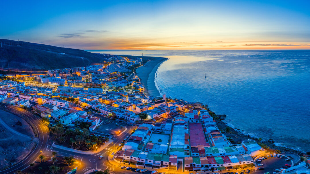 A picturesque 4K wallpaper showcasing an aerial view of Morro Jable Town and the expansive Playa del Matorral beach on Fuerteventura at dusk. The town's warm streetlights glow against the cool twilight sky, where a strip of orange sunset meets the deep blue Atlantic Ocean, creating a serene and vibrant coastal scene.