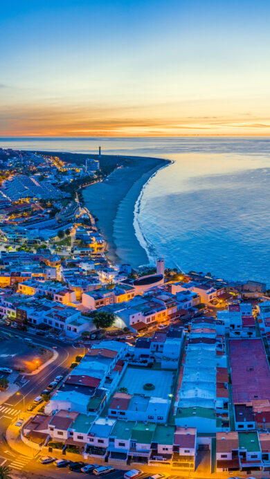 A picturesque 4K wallpaper showcasing an aerial view of Morro Jable Town and the expansive Playa del Matorral beach on Fuerteventura at dusk. The town's warm streetlights glow against the cool twilight sky, where a strip of orange sunset meets the deep blue Atlantic Ocean, creating a serene and vibrant coastal scene.