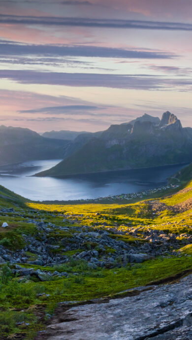 A breathtaking 4K wallpaper capturing the majestic Mount Segla on Senja Island, Norway, overlooking a serene fjord landscape at sunset. Golden sunrays burst over the distant peaks, casting warm light on the rugged green hillsides and reflecting soft hues on the tranquil water below.
