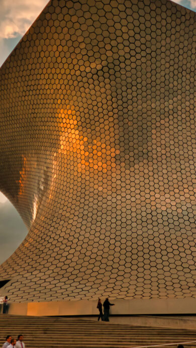 A breathtaking 4K wallpaper showcases the iconic Museo Soumaya at Plaza Carso in Mexico City, Mexico, under a dramatic sunset sky. The museum's shimmering, hexagonal facade glows with the warm, golden hues of the setting sun, creating a majestic and ethereal architectural scene.