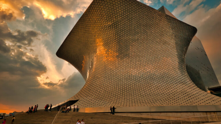 A breathtaking 4K wallpaper showcases the iconic Museo Soumaya at Plaza Carso in Mexico City, Mexico, under a dramatic sunset sky. The museum's shimmering, hexagonal facade glows with the warm, golden hues of the setting sun, creating a majestic and ethereal architectural scene.