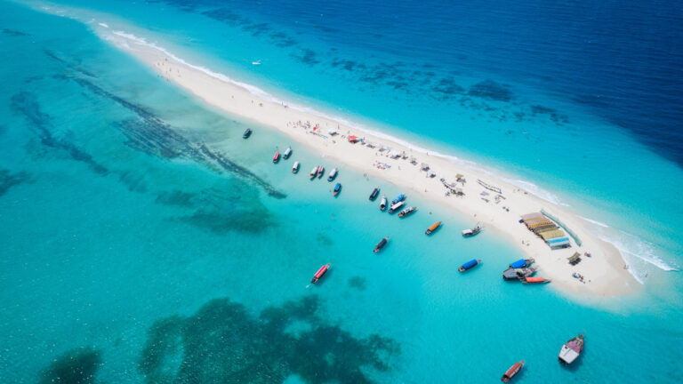 A breathtaking 4K wallpaper showcasing the Nakupenda Beach Sandbar from an aerial view in Zanzibar, Tanzania, where a thin strip of white sand is dotted with sunbathers and numerous colorful boats are anchored nearby. The vibrant turquoise and deep blue waters surrounding the sandbar reveal clear seabed details, creating an inviting and idyllic tropical paradise scene.